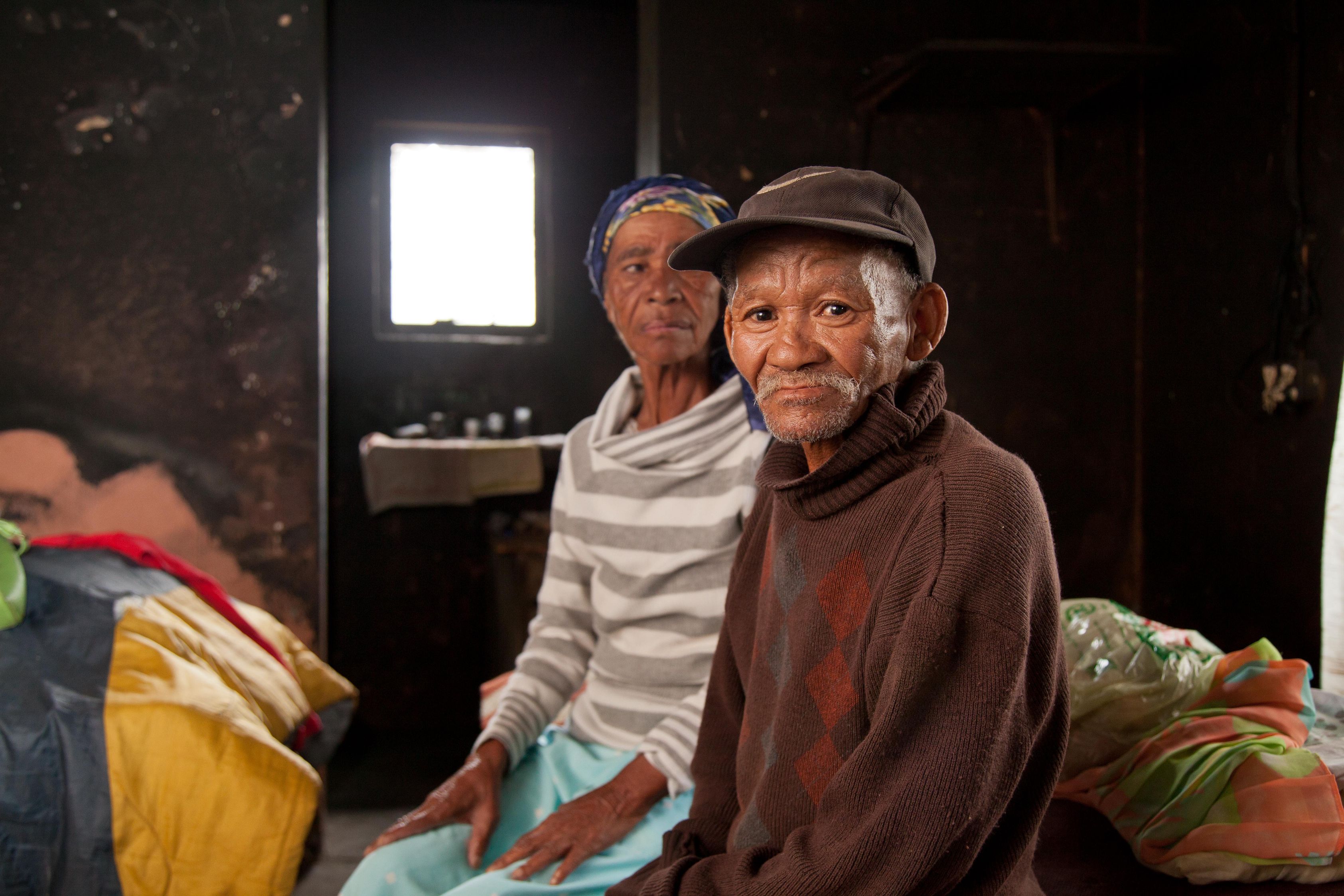 Ein älteres Paar Landarbeiter:innen sitzt im Inneren ihres Hauses. Der Mann schaut uns direkt an. Die Wände scheinen russgeschwärzt, man sieht Armut aber auch Liebe zum Heim.