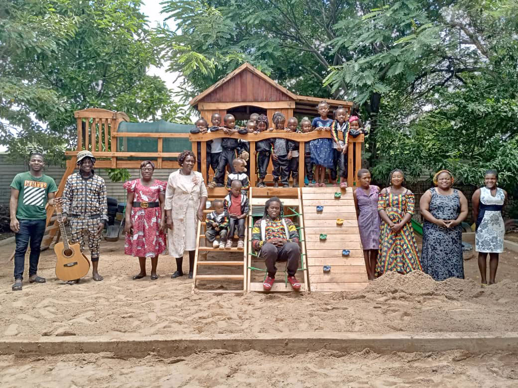Das hölzerne Spielhaus in der Vorschule von Dangwe mit Kindern und Betreuer:innen aufgereiht und in schönsten vielfarbigen Kleidern